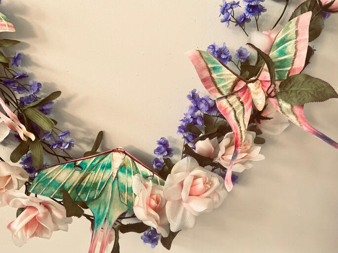 large pink and green moths on the same vine of pink roses. a few purple flowers hang behind them.